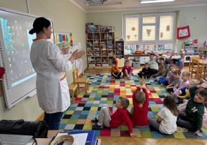 Dzieci siedzą w półkolu, a mama kolegi wita się z nimi i opowiada o czym będzie mówić, tj. pracy w aptece.