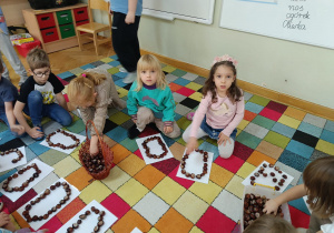 Dzieci siedzą na dywanie w kole i układają literkę „O” z kasztanów.