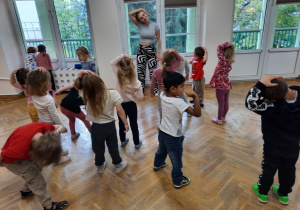 Dzieci stoją w rozypce i trzymając rękę na głowie lekko przyciągają ją do boku.