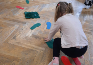 Dziewczynka stojąc na sensorycznych podkładkach stóp, kładzie ręce na sensoryczne podkładki rąk.