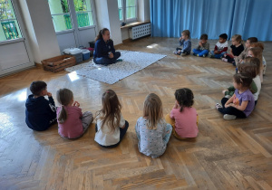 Dzieci siedzą w dużym półkolu na sali gimnastycznej, przed nimi na rozłożonym kocu siedzi Pani, za którą ułożone są różnej wielkości klatki dla zwierząt.