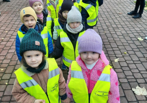 Maluchy ubrane w odblaskowe kamizelki stoją w parach przed wejściem do budynku przedszkola.