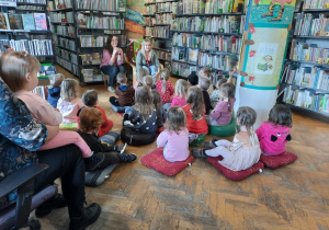 Przedszkolaki siedzą na poduszkach w sali biblioteki i słuchają Pani bibliotekarki.