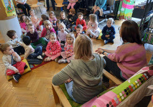 Przedszkolaki siedzą na poduszkach w sali biblioteki i słuchają Pani bibliotekarki.