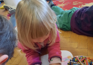 Dziewczynka leży na parkiecie i rysuje w serduszku prezent dla ukochanej osoby.