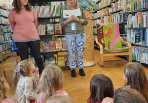 Dzieci siedzą na poduszkach i słuchają bajki czytanej przez Panią bibliotekarkę.