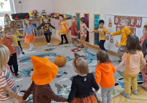 Dzieci poruszają się w dużym kole, trzymając się za ręce, do piosenki z okazji Dnia Dyni, przed nimi na środku dywanu leży duża dynia.