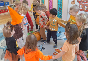 Przedszkolaki trzymając się za ręce zbliżają się do środka koła i stoją blisko dyni.