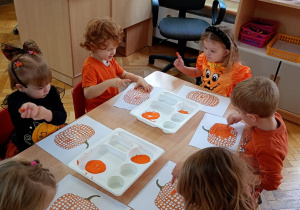 Dwóch chłopców i cztery dziewczynki siedzą przy stoliku, za pomocą paluszka stemplują na kartce wzór dyni.