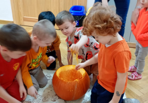 Na ceracie leży dynia z odciętym u góry ogonkiem, siedzący wokół niej chłopcy próbują wydrążyć jej środek za pomocą rączek.