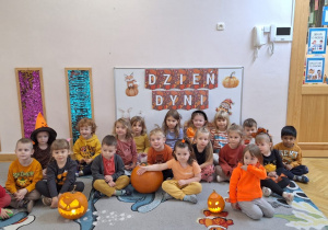 Cała grupa siedzi na dywanie na tle dekoracji z okazji Dnia Dyni, a przed sobą mają dwa dyniowe lampiony.
