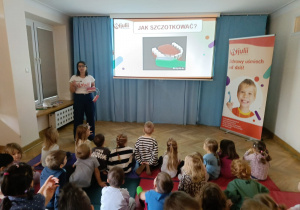 Przedszkolaki siedzą na materacach, obserwują jak prowadząca w oparciu o model szczęki i szczoteczki pokazuje im prawidłowy sposób szczotkowania zębów.