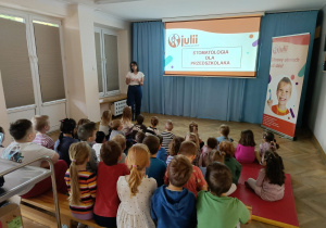 Dzieci siedzą na ławeczkach i materacach, przed nimi widać slajd pt. Stomatologia dla przedszkolaka, o którym opowiada prowdząca.