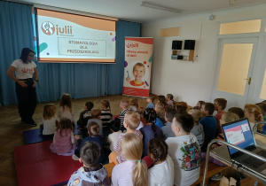 Dzieci siedzą na ławeczkach i materacach, przed nimi widać slajd pt. Stomatologia dla przedszkolaka, o którym opowiada prowdząca.