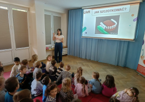 Dzieci siedzą na materacach, przed nimi widać slajd z pytaniem Jak szczotkować zęby, a prowadząca omawia to zagadnienie z użyciem szczoteczki.