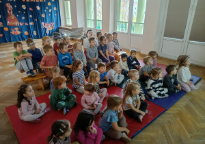 Przedszkolaki siedzą na materacach i ławeczkach słuchając prowadzącej.