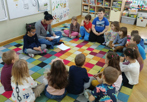 Dzieci siedzą w półkolu na dywanie i słuchają, jak mama jednej dziewczynki czyta wiersze w ramach projektu "Bohater Tygodnia".