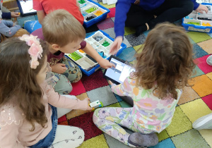 Dzieci siedzą na dywanie siedzą na dywanie i układają budowle z klocków Lego wg instrukcji wyświetlanej na tablecie na zajęciach z robotyki.