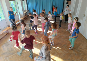 Dzieci stoją w rozsypce na sali gimnastycznej i powtarzają ruchy taneczne za trenerką zumby, podnosząc jedną nogę do góry.