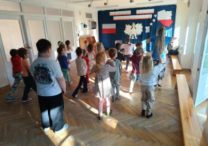 Dzieci stoją w rozsypce na sali gimnastycznej i powtarzają ruchy taneczne za trenerką zumby.