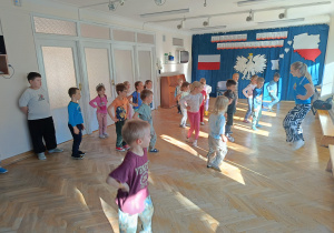 Dzieci stoją w rozsypce na sali gimnastycznej i powtarzają ruchy taneczne za trenerką zumby.