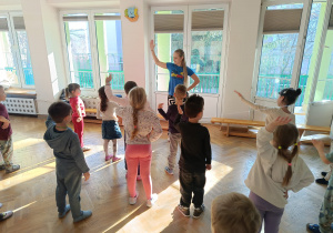 Dzieci stoją w rozsypce na sali gimnastycznej i powtarzają ruchy taneczne za trenerką zumby, trzymając rękę na boku, a drugą uniesioną w powietrzu.