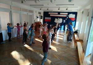 Dzieci stoją w rozsypce na sali gimnastycznej i powtarzają ruchy taneczne za trenerką zumby, machając rękami na boki.
