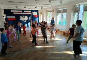 Dzieci stoją w rozsypce na sali gimnastycznej i powtarzają ruchy taneczne za trenerką zumby.