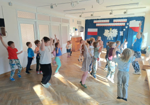 Dzieci stoją w rozsypce na sali gimnastycznej i powtarzają ruchy taneczne za trenerką zumby, podskakując do góry z podniesionymi rękami.