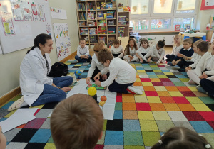 Dzieci siedzą na dywanie, a chłopiec z dziewczynką z pomocą prowadzącej odciskają foremki w piasku.