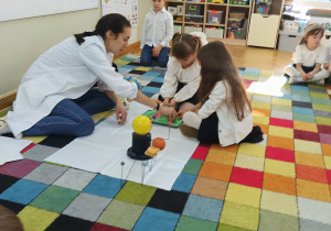 Dzieci siedzą na dywanie, a dwie dziewczynki z pomocą prowadzącej odciskają foremki w piasku.