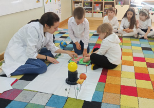 Dzieci siedzą na dywanie, a chłopiec z dziewczynką z pomocą prowadzącej odciskają foremki w piasku.