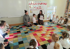 Dzieci siedzą na dywanie w półkolu, a chłopiec z mamą i ciocią prezentuje im wykonaną przez siebie książeczkę w ramach projektu Bohatera Tygodnia.