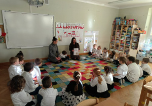 Dzieci siedzą na dywanie w półkolu, a chłopiec z mamą i ciocią prezentuje im wykonaną przez siebie książeczkę w ramach projektu Bohatera Tygodnia.