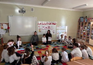 Dzieci siedzą na dywanie w półkolu, a chłopiec z mamą i ciocią prezentuje im kolorowanki z obrazkami z różnymi emocjami w ramach projektu Bohatera Tygodnia.