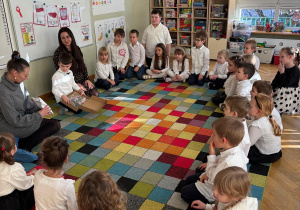 Dzieci siedzą w kole, a chłopiec z mamą i ciocią rozdają im małe upominki na zakończenie zajęć.