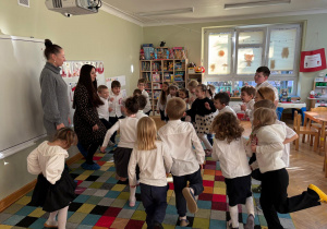 Dzieci stoją w rozsypce na dywanie i wspólnie z prowadzącymi wykonują różne ćwiczenia gimnastyczne stając na jednej nodze.