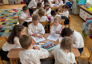 Dzieci siedzą przy stolikach i kolorują kolorowanki przedstawiające różne emocje.