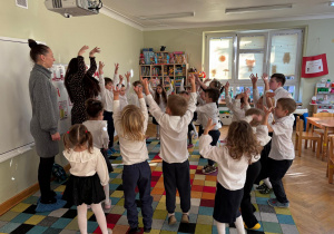 Dzieci stoją w rozsypce na dywanie i wspólnie z prowadzącymi wykonują różne ćwiczenia gimnastyczne, podnosząc ręce do góry.