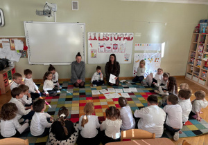 Dzieci siedzą na dywanie w półkolu, a chłopiec z mamą i ciocią prezentuje im kolorowanki z obrazkami z różnymi emocjami w ramach projektu Bohatera Tygodnia.