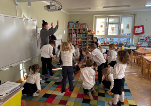 Dzieci stoją w rozsypce na dywanie i wspólnie z prowadzącymi wykonują różne ćwiczenia gimnastyczne, kucając.