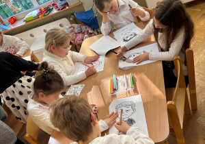 Dwóch chłopców i trzy dziewczynki siedzą przy stolikach i kolorują kolorowanki przedstawiające różne emocje.