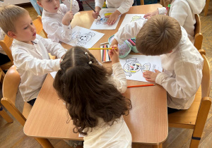 Dwie dziewczynki i czterech chłopców siedzi przy stolikach i koloruje kolorowanki przedstawiające różne emocje.