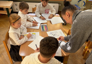 Trzech chłopców i dziewczynka siedzi siedzi przy stolikach i kolorują kolorowanki przedstawiające różne emocje.