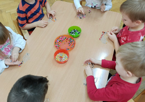 Dzieci siedzą przy stolikach i układają kolorowe koraliki na specjalnych plastikowych podkładkach.
