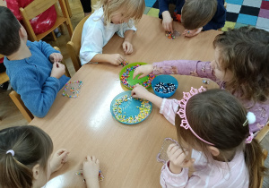 Dzieci siedzą przy stolikach i układają kolorowe koraliki na specjalnych plastikowych podkładkach.