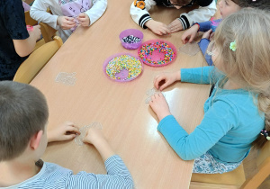 Dzieci siedzą przy stolikach i układają kolorowe koraliki na specjalnych plastikowych podkładkach.