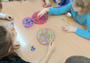 Dzieci siedzą przy stolikach i układają kolorowe koraliki na specjalnych plastikowych podkładkach.