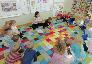 Dzieci siedzą w kole i wspólnie z koleżanką i jej mamą oglądają wykonane przez nich prace z kolorowych koralików.