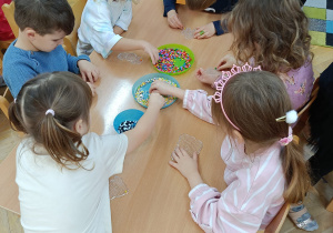 Dzieci siedzą przy stolikach i układają kolorowe koraliki na specjalnych plastikowych podkładkach.